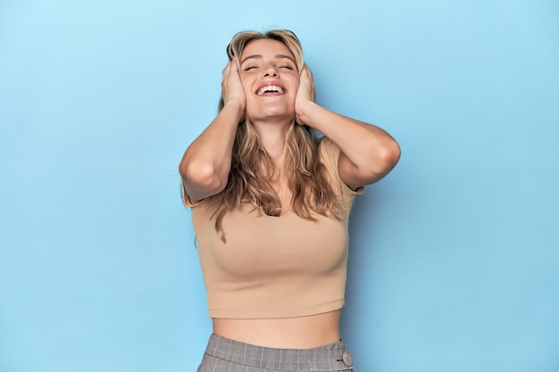 Une jeune femme blanche blonde dans un studio bleu rit joyeusement en gardant les mains sur la tête Concept de bonheur