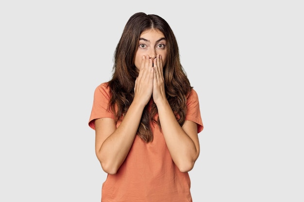 Jeune femme blanche dans le studio choquée couvrant la bouche avec des mains anxieuses