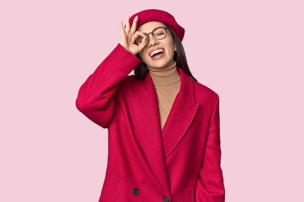Photo une jeune femme blanche élégante en trench-coat rouge et béret excitée en gardant le geste ok sur l'œil