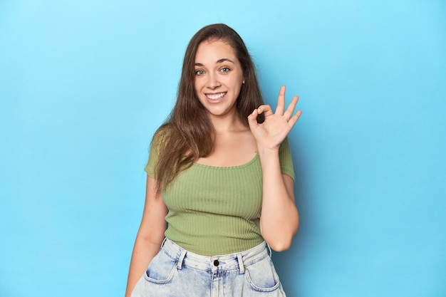 Jeune femme blanche en haut vert sur un fond bleu joyeuse et confiante montrant un geste ok