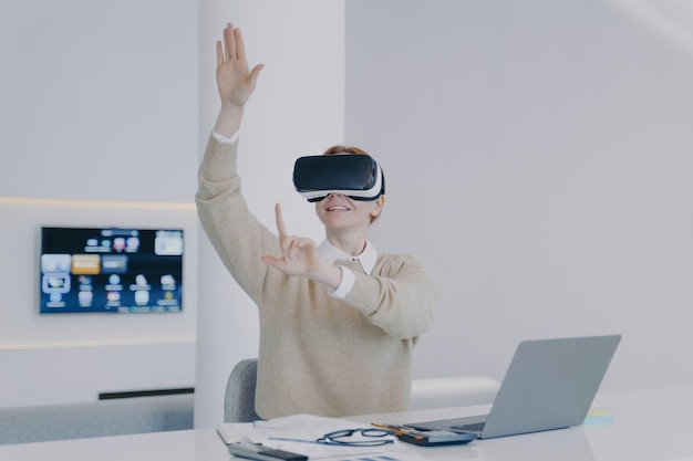 Jeune femme blanche en lunettes VR travaillant sur un projet dans le cyberespace devant un ordinateur portable au bureau