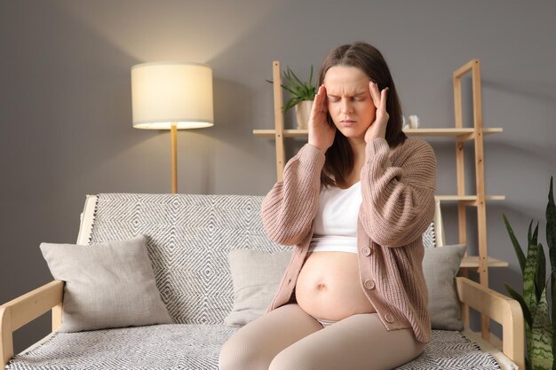 Une jeune femme blanche malade massant des tempes douloureuses faisant des grimaces de douleur assise sur le canapé portant des vêtements décontractés souffrant de migraines au troisième trimestre de grossesse