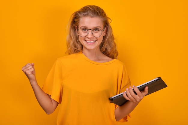 Une jeune femme blanche ravie avec une tablette à la main fait un geste victorieux.