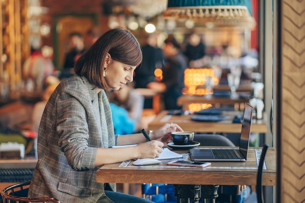Jeune femme en blazer avec ordinateur portable dans le café près de la fenêtre. Professions est blogueuse, pigiste et écrivain. Indépendant travaillant dans un café. Apprendre en ligne.
