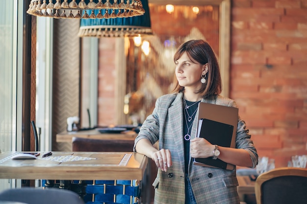 Jeune femme en blazer avec ordinateur portable et livre dans le café près de la fenêtre. Professions est blogueuse, pigiste et écrivain.