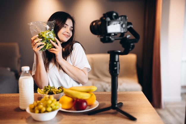 Jeune femme blogueuse alimentation saine cuisine fraîche de fruits salade végétalienne en studio de cuisine, tutoriel de tournage à la caméra pour chaîne vidéo. Une jeune influenceuse montre son amour pour une alimentation saine.