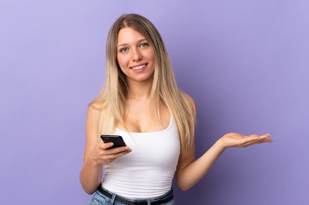 Jeune femme blonde à l'aide de téléphone mobile isolé sur violet tenant copyspace imaginaire sur la paume