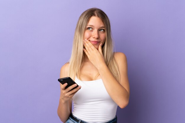 Jeune femme blonde à l'aide de téléphone portable isolé sur le mur violet en pensant à une idée