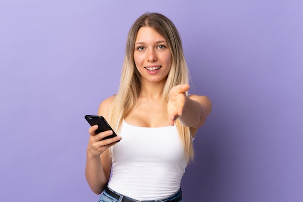 Jeune femme blonde à l'aide de téléphone portable isolé sur le mur violet poignée de main après une bonne affaire