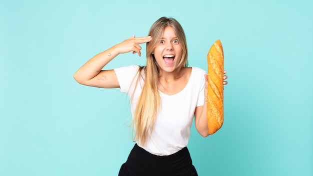 Jeune femme blonde à l'air malheureuse et stressée, geste de suicide faisant un signe d'arme à feu et tenant une baguette de pain