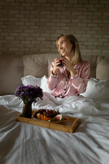 Jeune femme blonde appréciant le petit déjeuner au lit tôt le matin