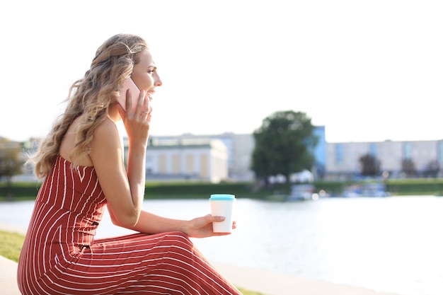 Jeune femme blonde assise au bord de la rivière à l'extérieur parlant par téléphone mobile.