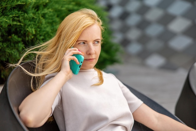 Jeune femme blonde assise à l'extérieur dans la ville et passant un appel avec son smartphone Fille parlant avec des amis avec un téléphone portable Communication d'activité de loisirs Conversation Technologie mobile