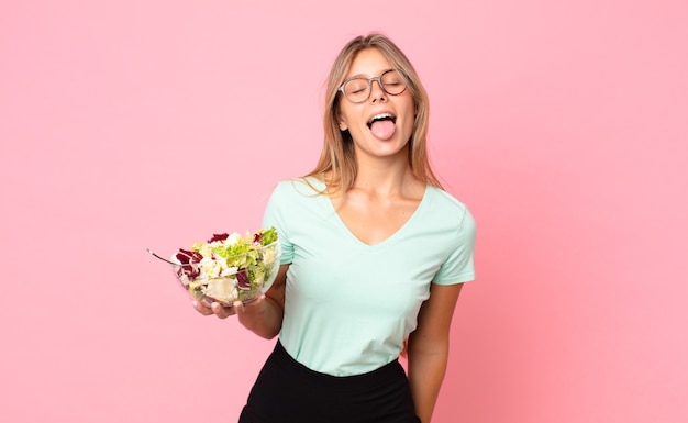 Jeune femme blonde à l'attitude joyeuse et rebelle, plaisantant et tirant la langue et tenant une salade