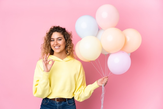 Jeune femme blonde aux cheveux bouclés attraper de nombreux ballons isolés sur rose montrant un signe ok avec les doigts