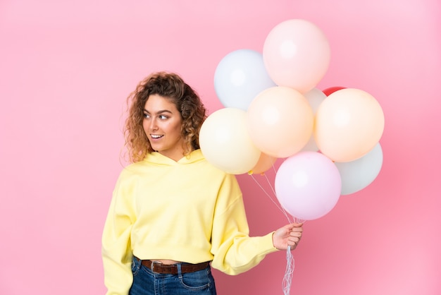 Jeune femme blonde aux cheveux bouclés attraper de nombreux ballons sur mur rose à la recherche sur le côté