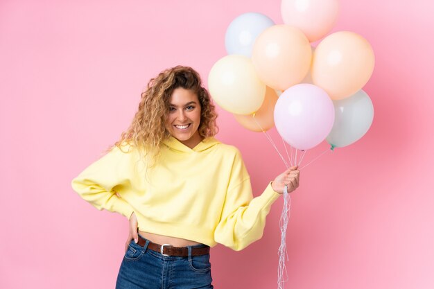 Jeune femme blonde aux cheveux bouclés attraper de nombreux ballons sur rose