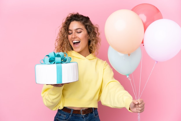 Jeune femme blonde aux cheveux bouclés attraper de nombreux ballons et tenant un gros gâteau isolé sur un mur rose