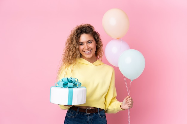 Jeune femme blonde aux cheveux bouclés attraper de nombreux ballons et tenant un gros gâteau sur le mur rose