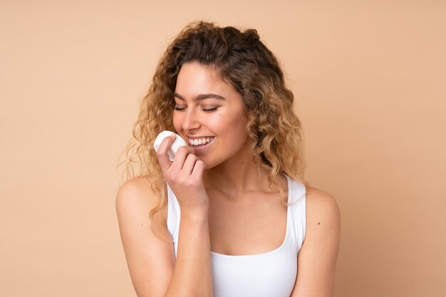 Jeune femme blonde aux cheveux bouclés isolé sur mur beige avec une crème hydratante et le sentir