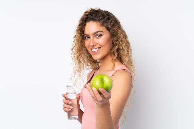 Jeune femme blonde aux cheveux bouclés isolé sur un mur blanc avec une pomme et avec une bouteille d'eau