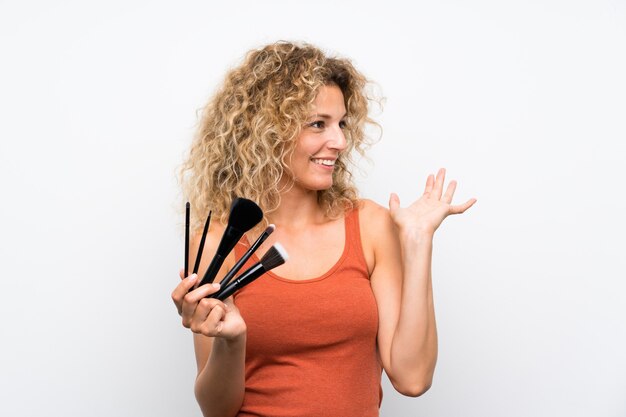 Jeune femme blonde aux cheveux bouclés tenant beaucoup de pinceau de maquillage avec une expression faciale surprise