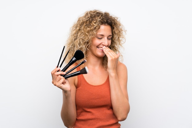 Jeune femme blonde aux cheveux bouclés tenant beaucoup de pinceau de maquillage souriant beaucoup