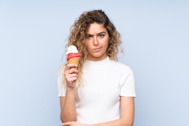 Jeune femme blonde aux cheveux bouclés tenant une glace au cornet sur bleu avec une expression triste