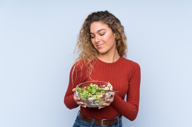 Jeune femme blonde aux cheveux bouclés tenant une salade sur mur isolé