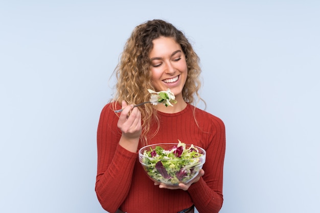 Jeune femme blonde aux cheveux bouclés tenant une salade sur le mur