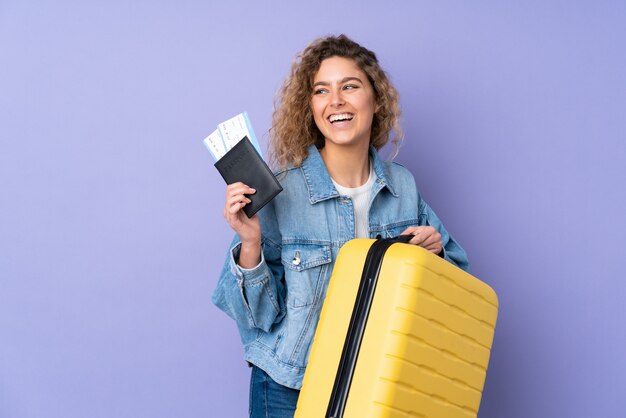 Jeune femme blonde aux cheveux bouclés sur violet en vacances avec valise et passeport