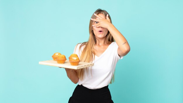 Jeune femme blonde ayant l'air choquée, effrayée ou terrifiée, couvrant le visage avec la main et tenant un muffins troy