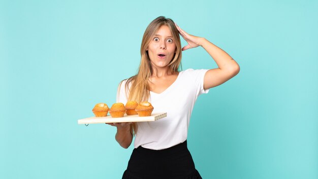 Jeune femme blonde ayant l'air heureuse, étonnée et surprise et tenant un muffins troy