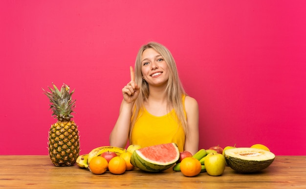 Jeune femme blonde avec beaucoup de fruits montrant avec l&#39;index une excellente idée