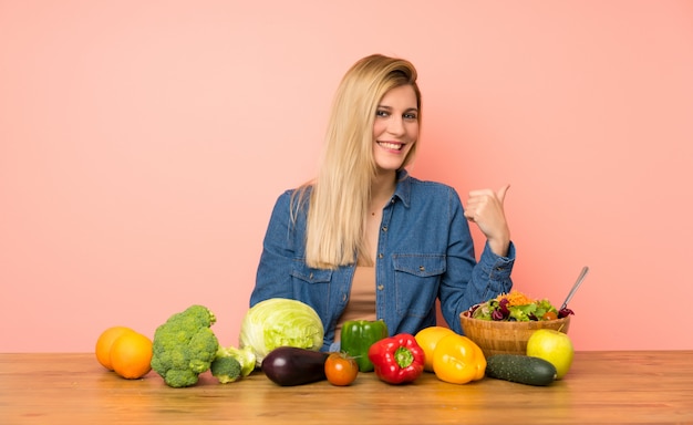 Jeune femme blonde avec beaucoup de légumes pointant sur le côté pour présenter un produit