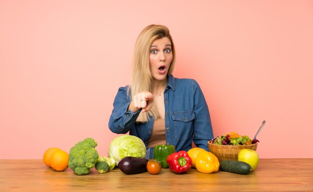 Jeune femme blonde avec beaucoup de légumes surprise et pointant devant