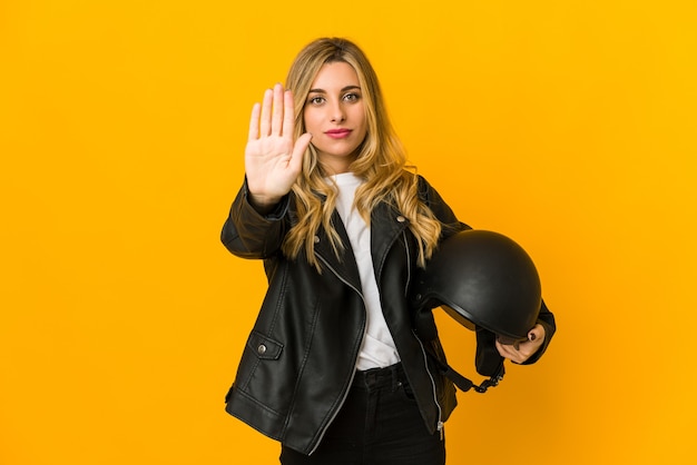 Jeune femme blonde biker caucasien tenant le casque debout avec la main tendue montrant le panneau d'arrêt, vous empêchant.