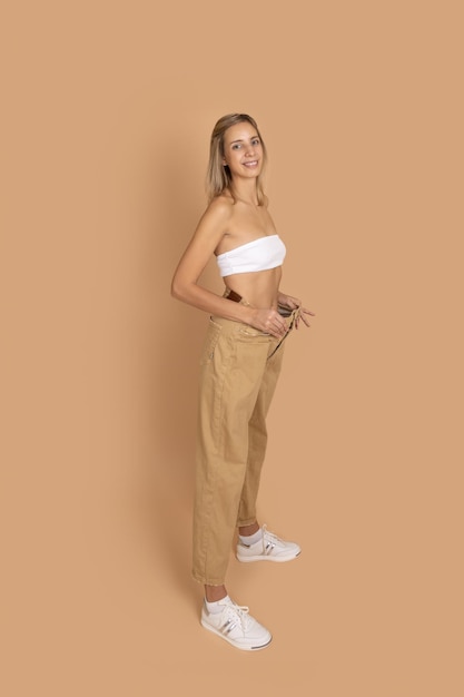 Jeune femme blonde en bonne forme souriante portant de gros pantalons surdimensionnés pour montrer la perte de poids sur fond de couleur peau Garder une alimentation saine faire de l'exercice