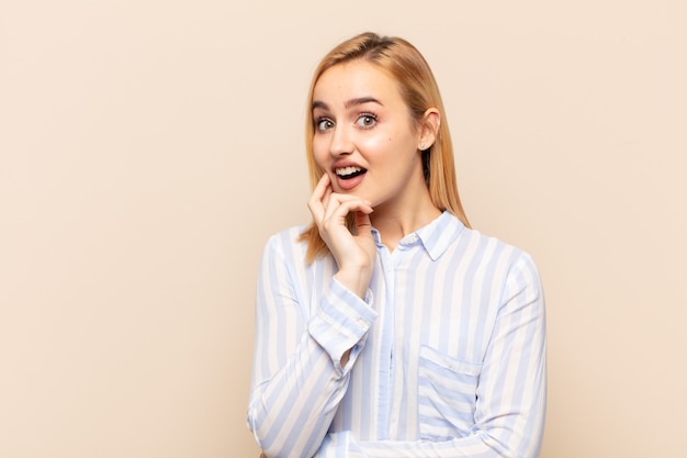 Jeune femme blonde bouche bée de choc et d'incrédulité, avec la main sur la joue et le bras croisé, se sentant stupéfaite et étonnée