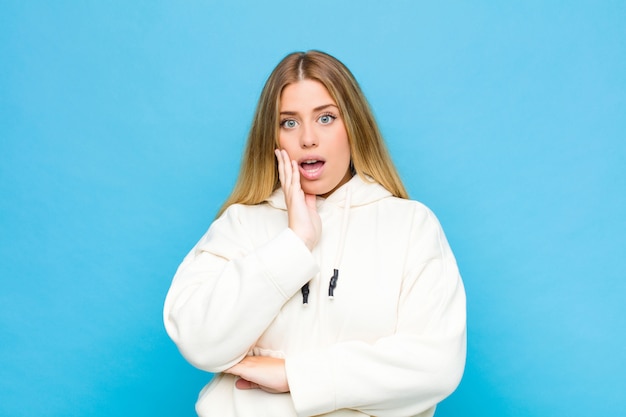 Jeune femme blonde bouche bée de choc et d'incrédulité, avec la main sur la joue et le bras croisés, se sentant stupéfaite et étonnée contre un mur plat