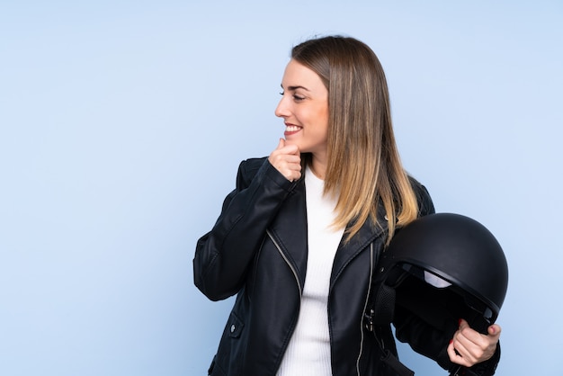 Jeune femme blonde avec un casque de moto sur un mur bleu isolé en pensant à une idée et à côté