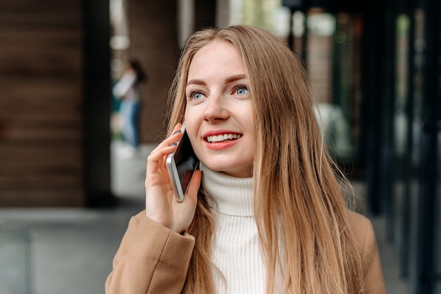 Jeune femme blonde caucasienne dans un manteau beige parlant sur un téléphone portable, souriant, tenant un dossier et regardant loin contre un immeuble de bureaux. Employé d'entreprise. Spécialiste en informatique