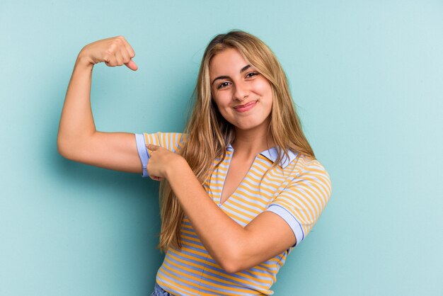 Jeune femme blonde caucasienne isolée sur fond bleu montrant un geste de force avec les bras, symbole du pouvoir féminin