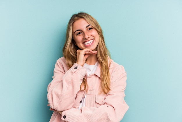 Jeune femme blonde caucasienne isolée sur fond bleu souriante heureuse et confiante, touchant le menton avec la main.