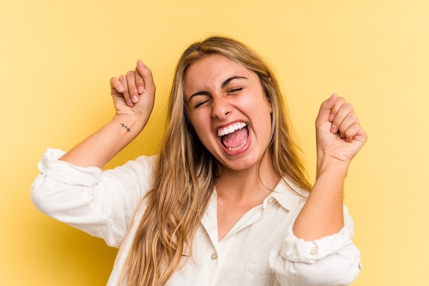 Jeune femme blonde caucasienne isolée sur fond jaune célébrant une journée spéciale, saute et lève les bras avec énergie.