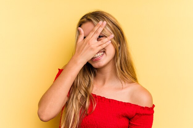 Jeune femme blonde caucasienne isolée sur fond jaune clignote à l'appareil photo à travers les doigts, gêné couvrant le visage.