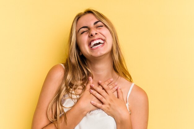 Jeune femme blonde caucasienne isolée sur fond jaune éclate de rire en gardant la main sur la poitrine.