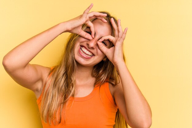 Jeune femme blonde caucasienne isolée sur fond jaune montrant un signe d'accord sur les yeux