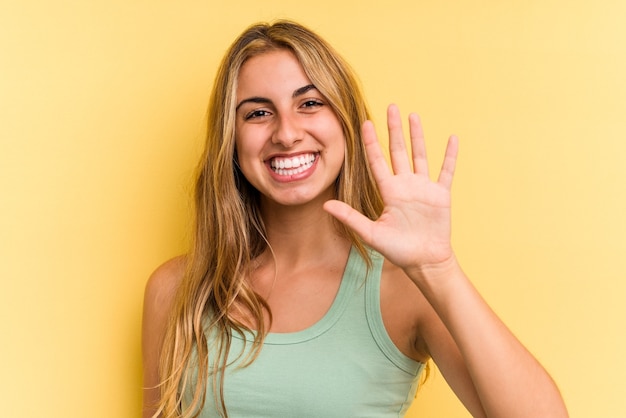 Jeune femme blonde caucasienne isolée sur fond jaune souriant joyeux montrant le numéro cinq avec les doigts.