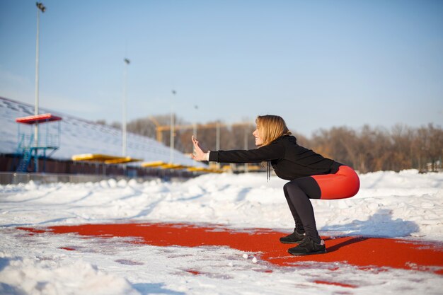 Jeune Femme Blonde Caucasienne En Legging Violet Exercice D'étirement Sur Une Piste De Course Rouge Dans Un Stade Enneigé. Fit Et Style De Vie Sportif